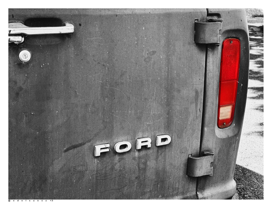 Schwarzweißfotografie mit einem farbigen Akzent: alter Ford Transporter aus den 1980er Jahren USA, im Bild das Heck mit schwarzmatt lackierter Tür, nur das hochkant stehende Rücklicht ist in leuchtendem Rot dargestellt, mittig der Ford Schriftzug in einzelnen Großbuchstaben