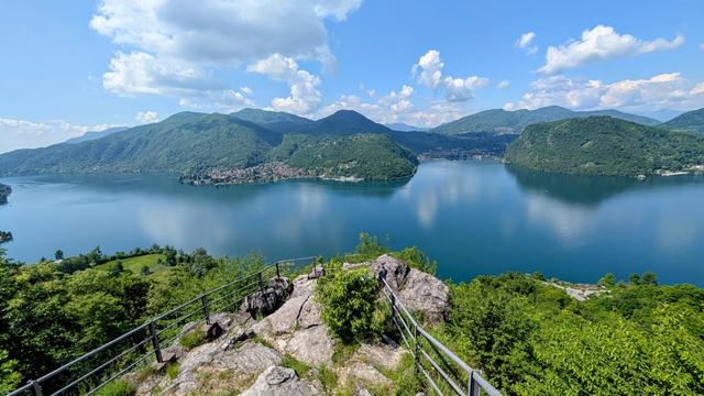 The image shows a stunning panoramic view of Lake Lugano (Lago di Lugano) from an elevated viewpoint with a metal railing. The calm blue waters of the lake are surrounded by lush green mountains under a bright blue sky with scattered white clouds. In the foreground is a rocky outcrop with a viewing platform, while the middle ground captures the serpentine shape of the lake winding between mountains. Small settlements can be seen nestled along the shoreline, and the overall scene represents a beautiful Alpine landscape on the border between Switzerland and Italy, photographed on a sunny day in spring.