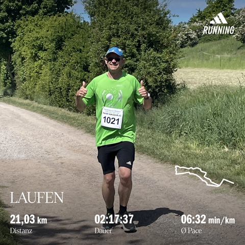 Läufer in grünem Shirt, mit einer Startnummer und mit schwarzer kurzer Laufhose und blauem Käppi laufend auf einem Feldweg mit zwei nach oben zeigenden Daumen, im Hintergrund grüne Wiese und Sträucher