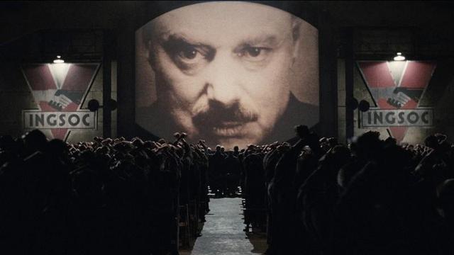 Sala de cine abarrotada de gente en las butacas y muy oscura mirando a una pantalla gigante con el rostro del Gran Hermano, y a ambos lados estandartes con el logo del Partido.