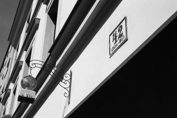 The black and white image shows the exterior of a building with a street sign that reads “42” and “Münzergasse” mounted above an archway, and a decorative wrought iron bracket holding a spherical warning lamp.