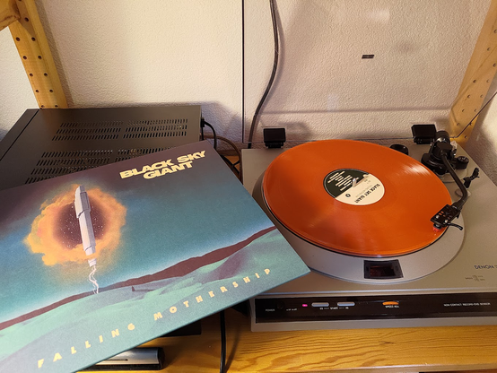 Black Sky Giant's "Falling Mothership" playing on a Denon DP-30L turntable. The cover shows a vertical spaceship (very similar to the mothership in the Homeworld series of games) hovering over a green desert planet, with a tiny figure beneath it. The sky behind the spaceship lookes like it has had a hole ripped in it, and stars shine through. The record itself is a deep rich orange.