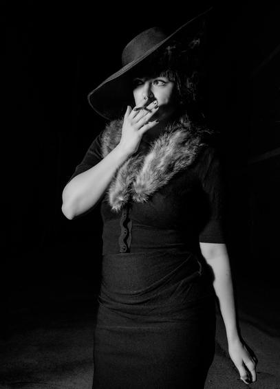 Black and white photo of a young woman smoking a cigarette in a dark alley. She's dressed as a femme fatale from an earlier era of movies, with a wide-brimmed hat and a fur-lined dress. She's looking cautiously to her right, as though danger might be approaching from an unseen corner.