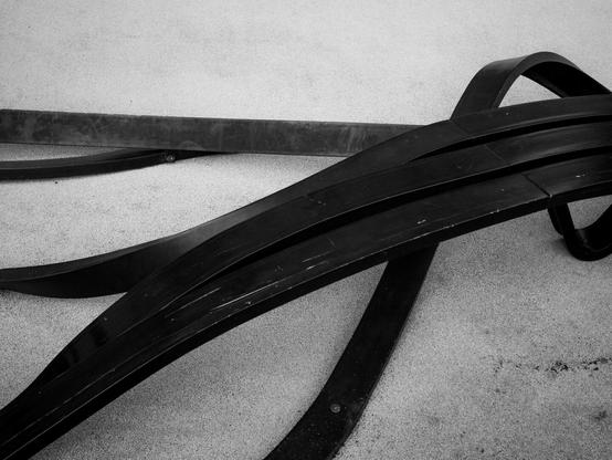 Monochrome photo of a black slat bench against a white background, curving in from the top right, already decohering as the slats bend and twist apart, to the extent that they exit the frame spread from the top left down to the middle of the bottom edge.