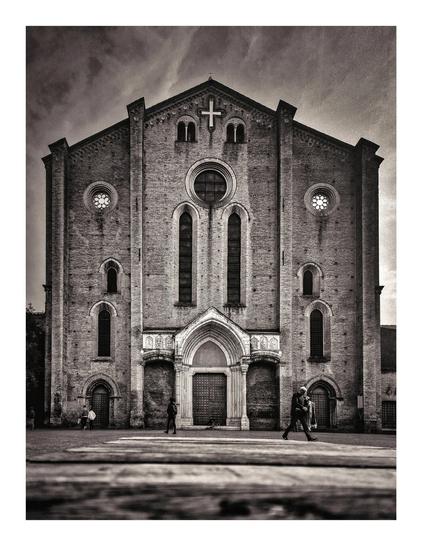 This is a black and white photograph depicting the facade of a grand, imposing brick church. (La Basilica di San Francesco) It Romanesque in style. There are arched windows and doors, and a large central rose window. The building is made of dark bricks, showing signs of age and weathering.  A cross is prominently featured at the top. The sky above the church is a little bit dark and cloudy.
In the foreground, there are a few people walking in front of the church. They look very small in comparison to the size of the basilica.