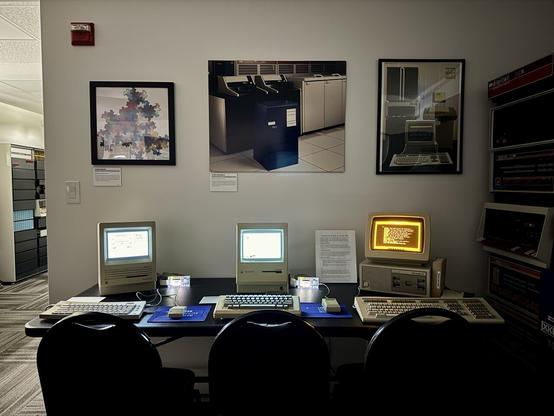 A Mac SE, Mac Plus and DECmate III on a table with various posters and artwork.