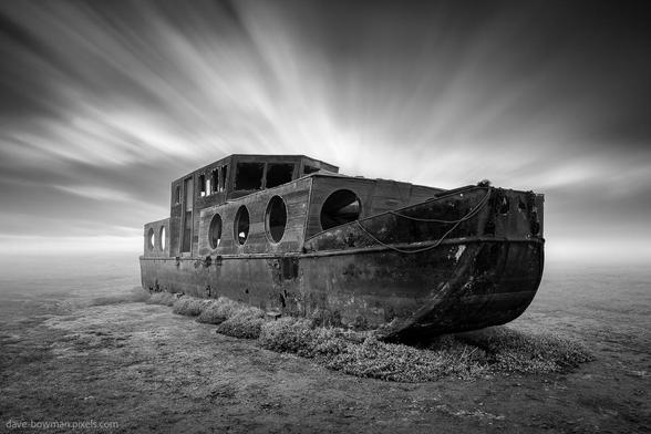 A photograph of an abandoned boat resting on the salt marshes at Blakeney, Norfolk, its worn frame isolated in the open landscape. The long exposure captures streaks of cloud sweeping across the sky, introducing gentle motion to the otherwise still scene. The black and white tones highlight the textures of the boat and land, giving the image a quiet, weathered character with a sense of time slowly unfolding.