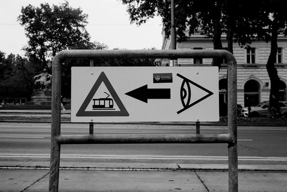The black and white image shows a street sign mounted on a metal frame, featuring three symbols: a tram inside a warning triangle, a bold left-pointing arrow, and a stylized eye, suggesting caution and awareness for trams coming from the left.
