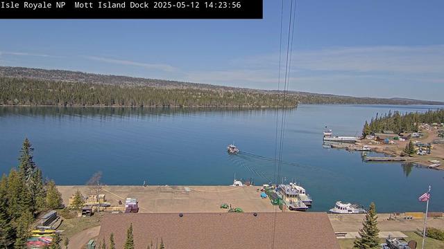 Boats in the water in this view from Isle Royale National Park today. In the foreground, a busy dock, with vessels coming and going. A tree-covered landscape fills the background. The surface of the water shows ripples from the movement of the boat and, at a larger scale, perhaps the energy of the wind. Near the far shore, the water is still enough to show a reflection of the trees.