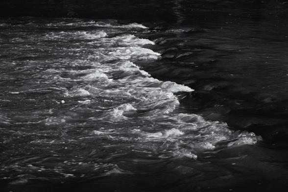 A black and white image of a ripple with bright white highlights against darker water. Simple composition.