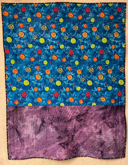 Back of the quilt. Top two thirds is a print on a blue background with bright swirling circles. The bottom seems to be a hand dyed purple.
