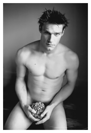a nude, fit man with spiky dark hair sits on a wooden chest cradling a dried hydrangea flower in his lap, making eye contact.