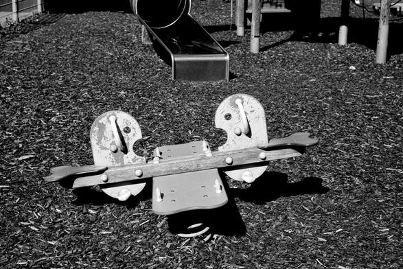 The black and white image shows an empty children’s playground with a worn, two-seat spring rocker in the foreground and a metal slide in the background, surrounded by wood chip ground cover.