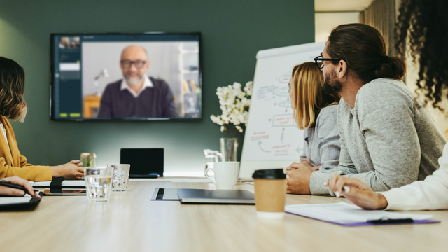 Teilnehmende einer Besprechung sitzen in einem Konferenzraum und verfolgen eine OpenTalk Videokonferenz auf einem großen Bildschirm. Auf dem Tisch liegen Laptops, Notizbücher und Getränke, im Hintergrund steht ein Flipchart mit Skizzen.