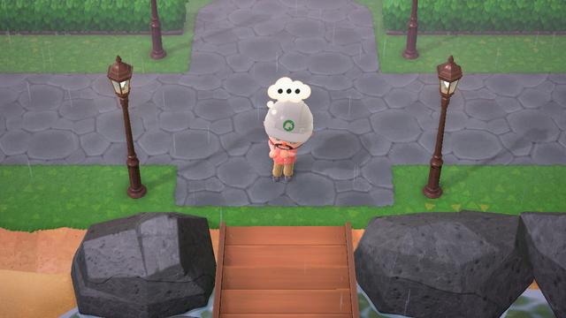 Animal Crossing: New Horizons screenshot set in the rain. A human villager seen from above wearing a white construction hat is standing at the entrance which has a concrete walkway in front of them and going in all directions away. On either side of the entrance are copper colored streetlamps. Another set of them can be partially seen behind the villager at an entrance between some partially visible hedge fences.