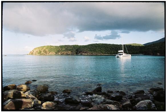 A film photograph of a cove with azure water. There is a rocky shore in the foreground and a green vegetated point of land in the background. A catamaran sailboat is anchored in the cove.