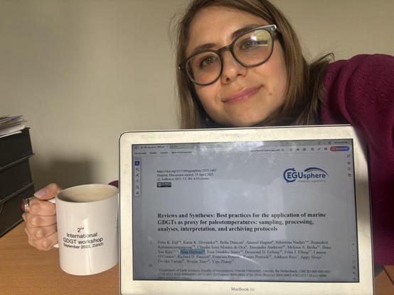 Nina Davtian (that's me!) at her work office, with the laptop screen showing the header of her co-authored preprint by Bijl and Sliwinska et al. (submitted to Biogeosciences) EGUsphere (https://doi.org/10.5194/egusphere-2025-1467) with her name highlighted in the author list. Nina is holding a mug on which the text reads « 2nd international GDGT workshop September 2023, Zürich ».