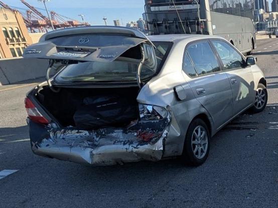 A very sad early Prius that's all smashed up on the highway. It has a visibly crumpled rear end and an open trunk.