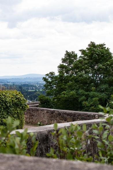 Eine Steinmauer und üppiges Grün überblicken eine ferne Landschaft unter bewölktem Himmel und zeigen sanfte Hügel und Bäume.