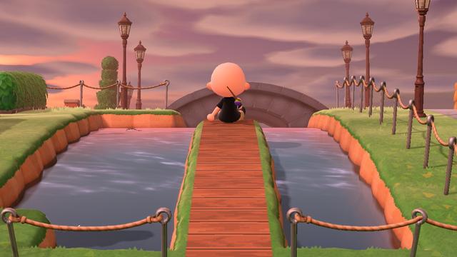 Animal Crossing: New Horizons screenshot. A bald, human villager with a fishing pole over their shoulder is sitting on the end of a faux wooden dock out into a small water area. In the distance is a stone bridge and a line of street lamps on either side.