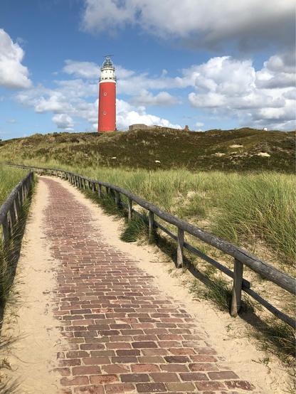 Das Bild zeigt den markanten roten Leuchtturm „Vuurtoren Eierland“ auf der niederländischen Nordseeinsel Texel. Der Leuchtturm steht auf einer kleinen Anhöhe inmitten von Dünenlandschaft. Der rote Turm mit weißem Kopf und Laterne hebt sich deutlich vom bewachsenen Hügel und dem teils bewölkten, aber blauen Himmel ab.

Im Vordergrund führt ein gepflasterter Weg aus roten Backsteinen, flankiert von einem einfachen Holzgeländer, durch den sandigen Dünensand direkt auf den Leuchtturm zu. Links und rechts des Weges wachsen typische Dünengräser und kleinere Büsche.