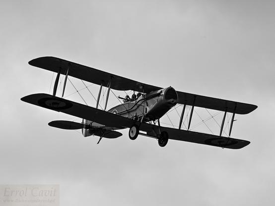 A two seat single engine biplane from its front quarter and below, solid grey cloud beyond. In a traditional 4:3 aspect and black and white.