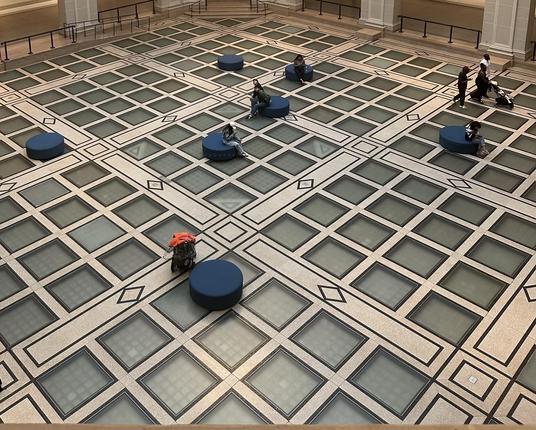 An aerial view of a spacious indoor area featuring a patterned tiled floor with glass panels and geometric designs. Several blue circular seating arrangements are scattered throughout, with a few people sitting and others walking, including someone with a stroller.