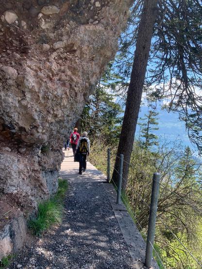 ein schmaker weg unter markanten nagelfluhfelsen. rechts mit drahtzaun gesichert. rückenansicht von zwei wandererinnen mit rucksack.