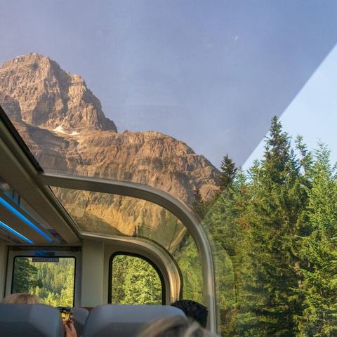 The image shows a prominent, rocky slate-brown mountain with a large patch of white snow, surrounded by a landscape of dense evergreen forest, viewed from inside a train with a glass dome observation car.