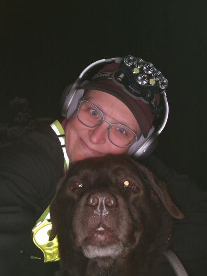 Night time selfie of a person and a chocolate brown Labrador with greying snout. The person is wearing a black jacket, yellow safety harness, red cap, silver headphones and blue-rimmed glasses. On their cap rests a headstrap torch in off position. Their free hand is around the dog's shoulders. Both are facing the camera. The person's head is resting on the dog's head. They're smiling, grateful for their time spent together.