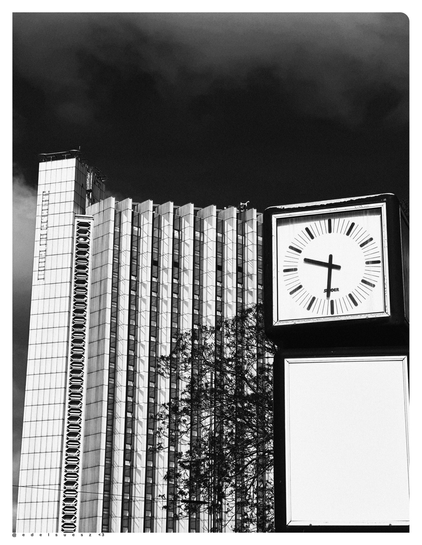 Schwarzweißfotografie: links ein helles eckiges Hochhaus, rechts auf einem Mast eine analoge Uhr, diese zeigt 9:31, darüber dunkler Himmel