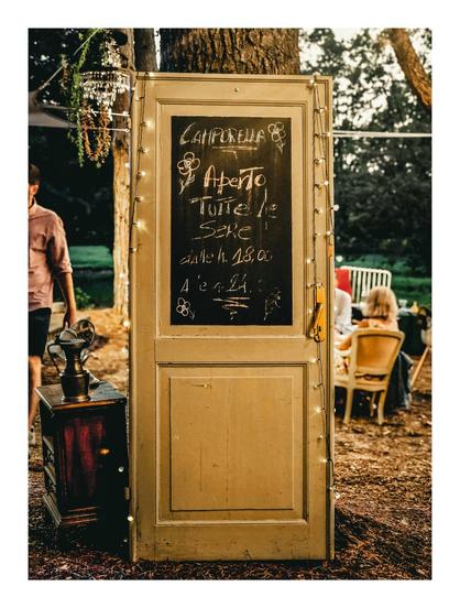 The photo is of an old, weathered light coloured door. It is outside and is leaning up against a tree. There are fairy lights around the outside edges of the door and the top frame of the door is a chalkboard. In the inside it displays "Camporella" the name of the bar and also says the opening times are from 6pm to 12am every evening. Behind the door there are blurred figures sitting and standing and you can see some of the rustic furniture of the bar.