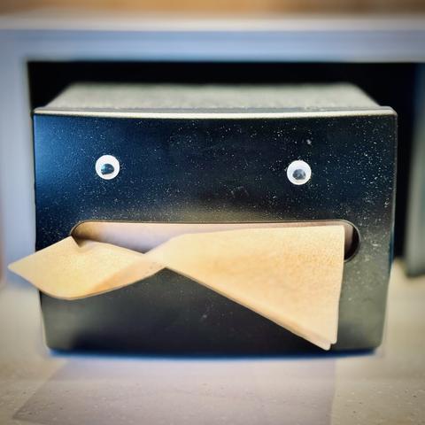 Googly eyes are glued to a commercial napkin dispenser, as a brown paper napkin hangs out of its center, resembling a mouth with a large tongue.