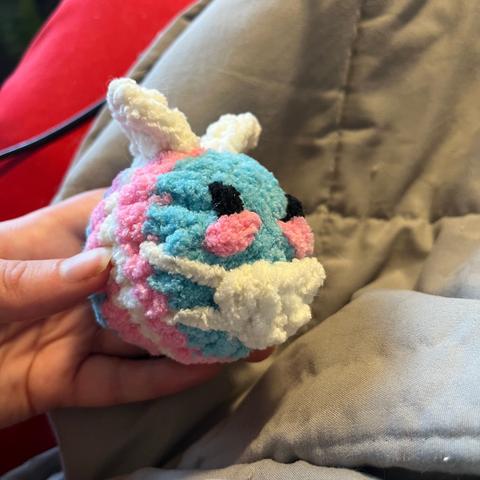 A hand holding a fuzzy bee knitted out of chenille yarn, with trans flag stripes and a knitted mask