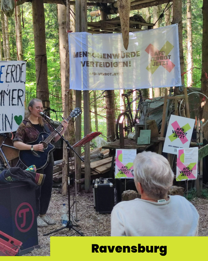 Ein Foto der Kundgebung in Ravensburg. Eine Person mit Gitarre steht vor dem Mikrophon und singt. Umgeben ist sie von Bäumen, einem Baumhaus und AfD-Verbotsschildern. Zwischen den Bäumen hängen Banner. Eine zuhörende Person ist von hinten zu sehen.