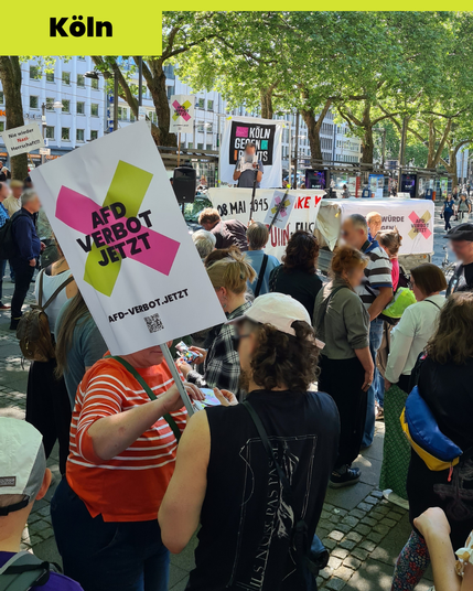 Ein Foto der Kundgebung in Köln. Ein Mensch hält gerade eine Rede. Er steht vor dem Banner "Köln gegen Rechts". Vor der Bühne sind mehrere Banner und Menschen mit Schildern. Eines der Schulder zeigt groß das Logo der Kampagne und den Link zur Webseite afd-verbot.jetzt