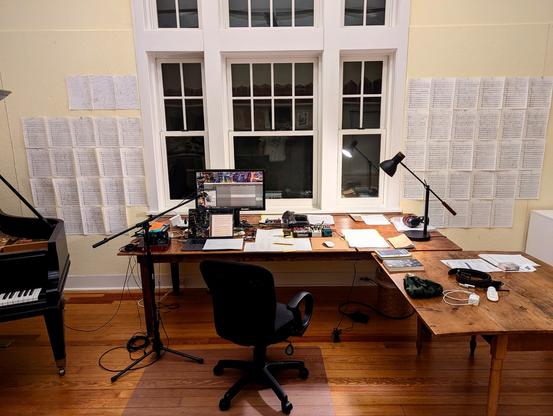 My studio during my stay at MacDowell. A large desk props my working space: laptop, monitor screen, audio interface, stompboxes, wiring, manuscript papers, books, a lamp. A microphone and a mic stand are set up. An additional desk on the right side has more books and other things. Cork walls on both sides of the large desk are filled with pinned manuscript paper I posted for inspiration. A baby grand piano on the left is partially visible in the photo.