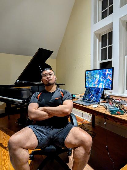 Me sitting in my chair at my desk, my arms folded in front of my chest. A baby grand piano, a microphone, and a mic stand are set up behind me. My workstation with my laptop, monitor screen, and other electronic gear are propped on the right.