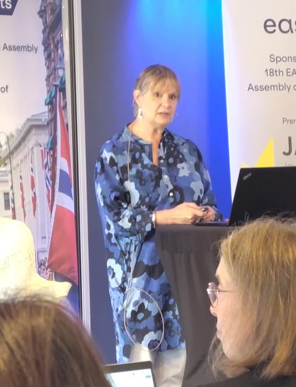 Alice Meadows at the EASE conference podium. She's a white woman with short greying hair, wearing earrings and a long blue patterned shirt.l and white trousers.