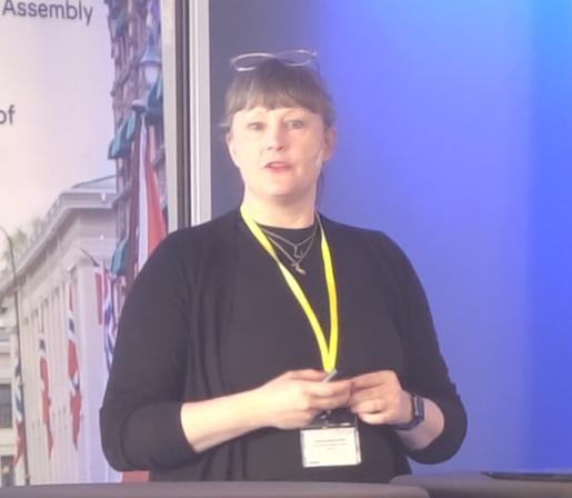 Sofie Wennström at the EASE conference podium. She's a white woman with black hair with bangs, wearing a black long sleeved top and with glasses on her head.