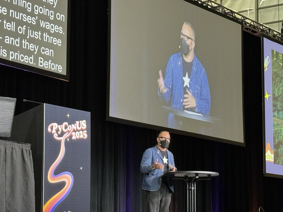 Cory Doctorow on stage at PyCon. He’s standing in front of a large screen with his image on it. To the left there is a podium with the PyCon US 2025 logo and another big screen with captions.