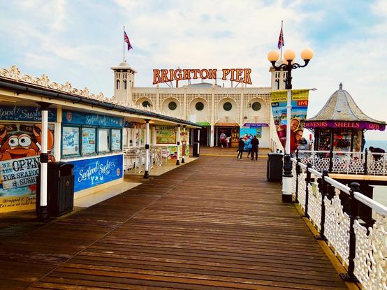 This image shows the Brighton Pier, a popular tourist destination in Brighton, England. The pier features various attractions and amenities, including food stalls, shops, and amusement facilities. Here, you can see a few businesses advertising seafood and sushi, along with other souvenir and gift shops. The architecture of the pier is prominently visible with its ornate details and festive decorations. Several people are casually walking along the pier, enjoying the environment. Flags are flown atop the structures, adding to the lively, welcoming atmosphere typical of such seaside piers.