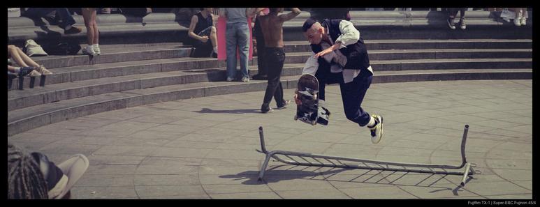 A skateboarder executes a trick jump over a fallen barrier in front of people seated on the 'steps' of a dried fountain.