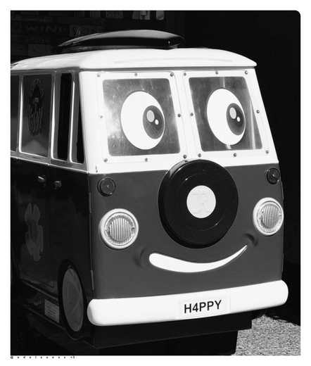 Schwarzweißfotografie: ein freundlich aussehender Spielzeugbus für Kinder in einer Fußgängerzone, fotografiert von schräg vorn, dieser ist dem VW Fensterbus nachempfunden, geteilte Frontfenster mit aufgemalten großen Augen, zwischen den stilisierten runden Frontscheinwerfern ein angedeutetes Reserverad darunter ein lachender Mund, darunter eine helle Stoßstange mit H4PPY als Nummernschild