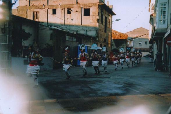 Analogic photo of a Galician street. The floor is made of big rectangular stone slabs, and appears a bit wet, as it was rainy that morning. In the moment of the picture the sun appeared, lighting up the upper part of the houses, but the street itself is mostly dark. There are some houses in the background and an old abandonded stone house with no windows at the left. Strings with little lanterns, attached to the houses, cross the street, signaling that we were celebrating festivities. In this case, Entroido (Galician carnival).

A line of more than a dozen people appears running from the background to the front left of the shot. The first person is a bit blurry. They're wearing the traditional Entroido costume from the town (cigarrón outfit): white pants and socks, a colorful jacket and a belt with cowbells around the waist, also a big wooden mask with a smiley face and moustache. Each cigarrón is carrying a small stick with a whip. Over their mask each participant wears a hat with a painted image of an animal, this picture is taken from certain distance and they don't appear clearly, but there were wolves, dogs, horses, eagles...