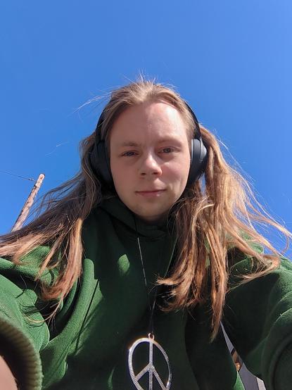 The image is a selfie taken by a young adult, in their mid twenties. They are wearing headphones and a green hoodie with a peace sign necklace. The background shows a clear blue sky and a light pole visible to the left.
