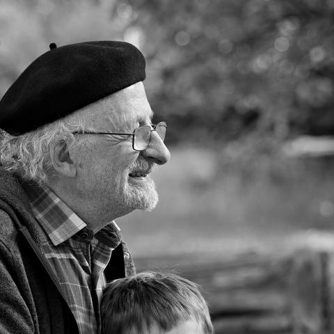 Quadratisches Schwarzweißbild, eines älteren Herren im Profil. Er trägt eine Baskenmütze unter der hinten leicht gewelltes helles Haar hervor kommt. Hinter der Brille sind Lachfalten um das Auge zu erkennen, der Mund ist vom Anflug des Lächelns leicht geöffnet.
Unten vor der Brust ist noch der Haaransatz einens Kindes zu sehen, das auf seinem Schoß sitzt.