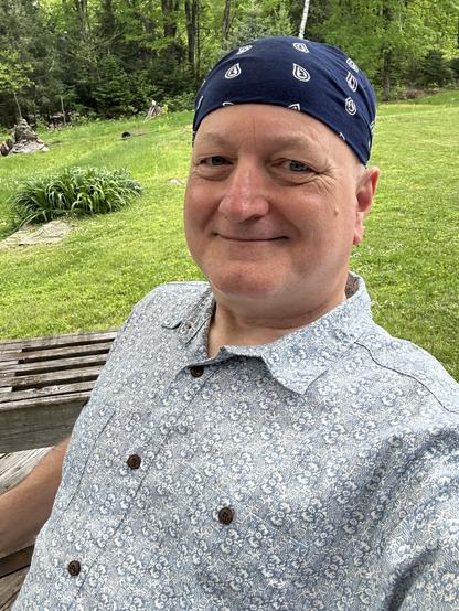 A smiling person with just some eyebrows for facial hair, wearing a dark blue head kerchief and a light blue and white patterned hemp and cotton button-up shirt. I am sitting on the porch with grass and trees behind me on a sunny day.