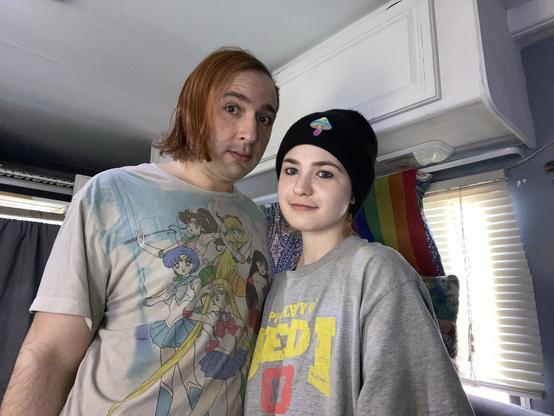 A person with shoulder-length reddish hair wearing a graphic t-shirt stands beside a person in a black beanie and oversized gray sweatshirt. The background features a rainbow flag and bright indoor lighting.