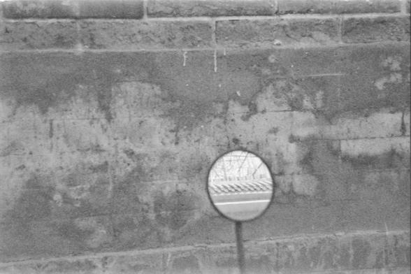 ### Image 1:
**English:**
A black and white photograph of a brick wall with a convex mirror attached to it. The mirror reflects a street scene, including a portion of a road and some indistinct buildings. The wall shows signs of wear and aging, with visible cracks and discoloration.

**Chinese:**
一张黑白照片，展示了一面砖墙，墙上安装有一个凸面镜。镜子反射出街道的景象，包括部分道路和一些模糊的建筑物。墙面显示出磨损和老化的迹象，有明显的裂缝和变色。